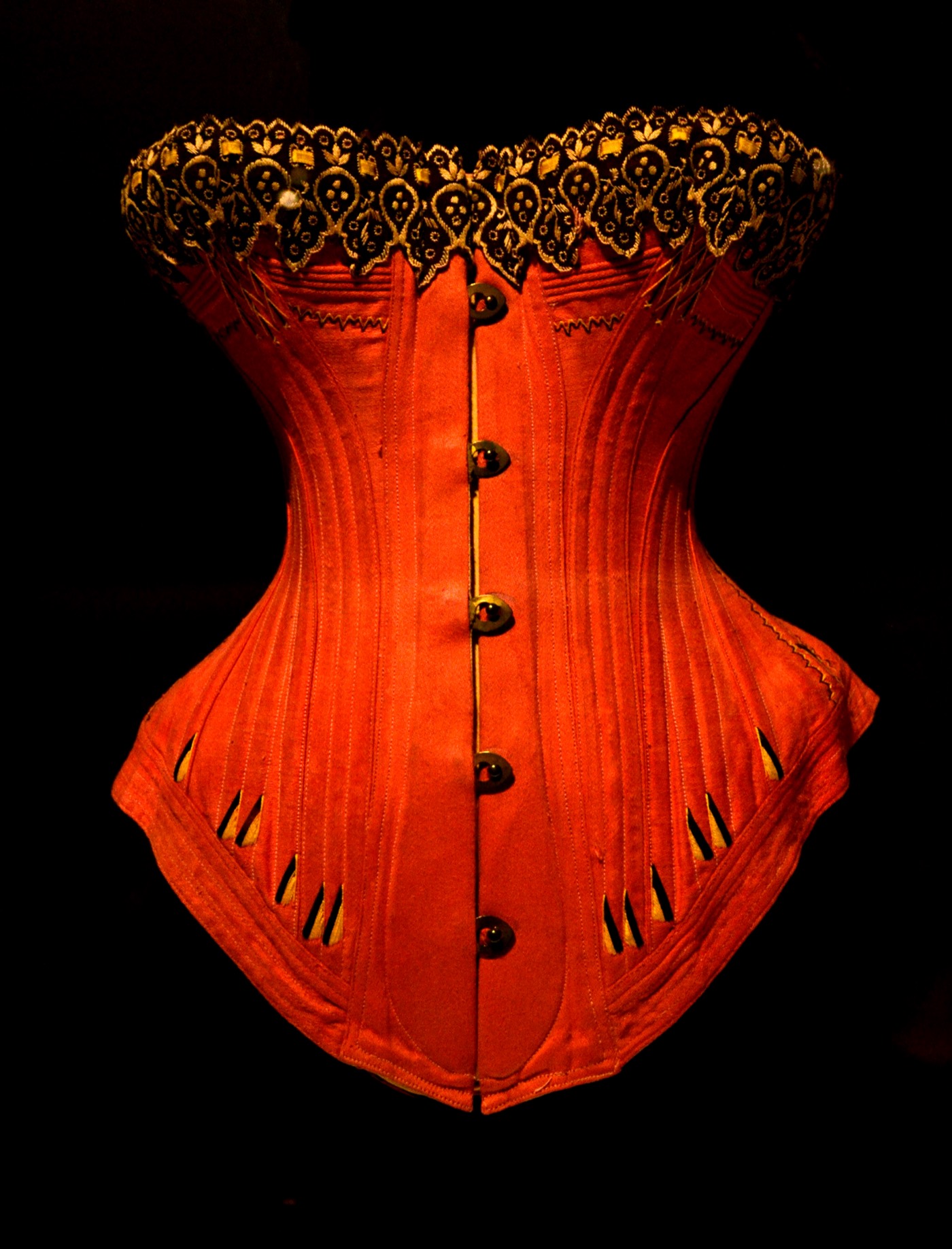 Front of an 1880s red satin corset. A two-part steel busk runs down the centre front, closed by five studs; boning channels run vertically through the panels, with flossing worked at the bone ends over the hips.